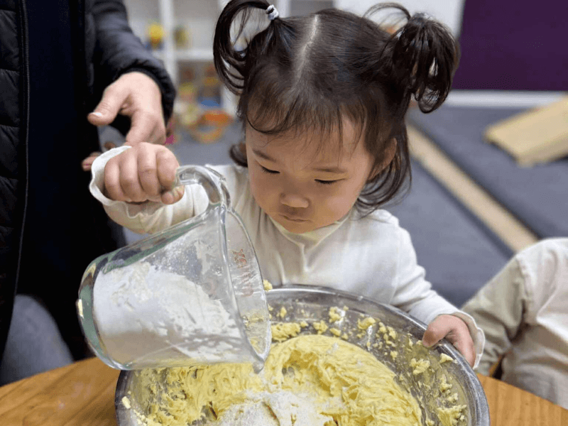 KidsCo pouring flour for playdough 800x600_compressed A young child exploring and pouring ingredients during a hands-on cooking activity.